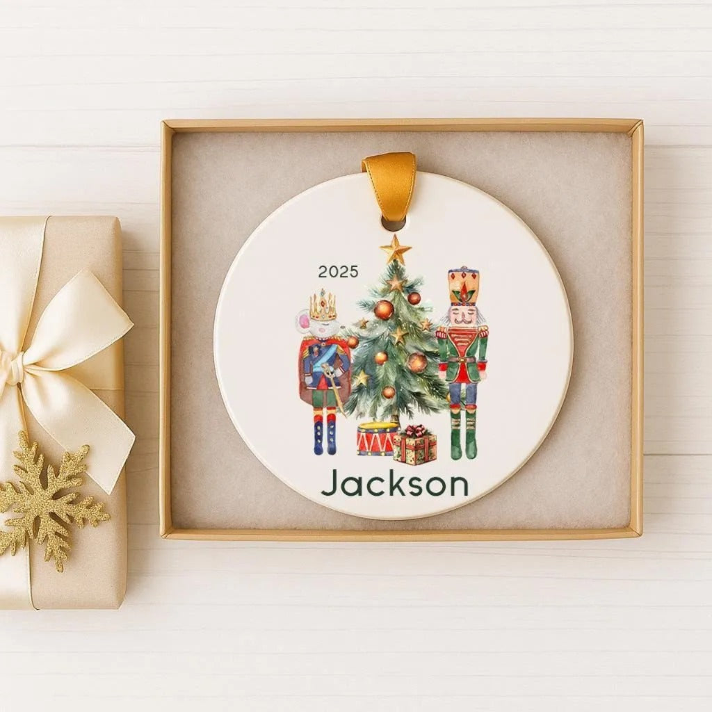 A white ceramic ornament with a Christmas tree and nutcracker figures, along with the name "Jackson" written on it, is displayed in a gold box.