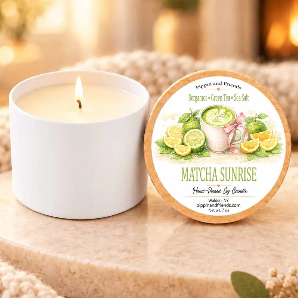 a white candle with a label that reads "MATCHA SUNRISE" and "Pippin and Friends" on a wooden surface, with a blurred background that includes a fireplace and some greenery.