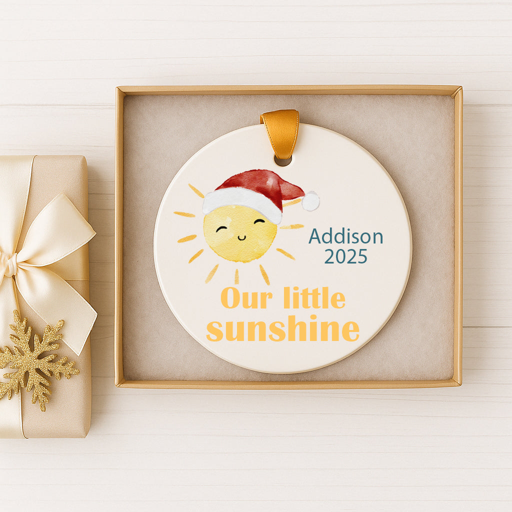 A white ornament with a smiling sun and a Santa hat, placed in a box with a gold ribbon.