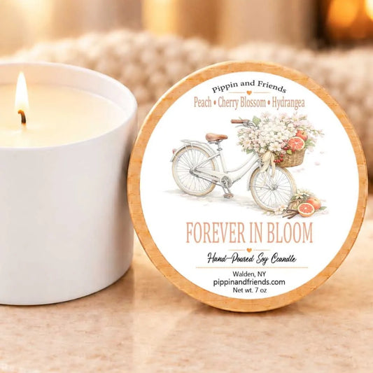 a white candle with a label that reads "Forever in Bloom" and a bicycle with a basket of flowers, placed on a table next to a lit candle.