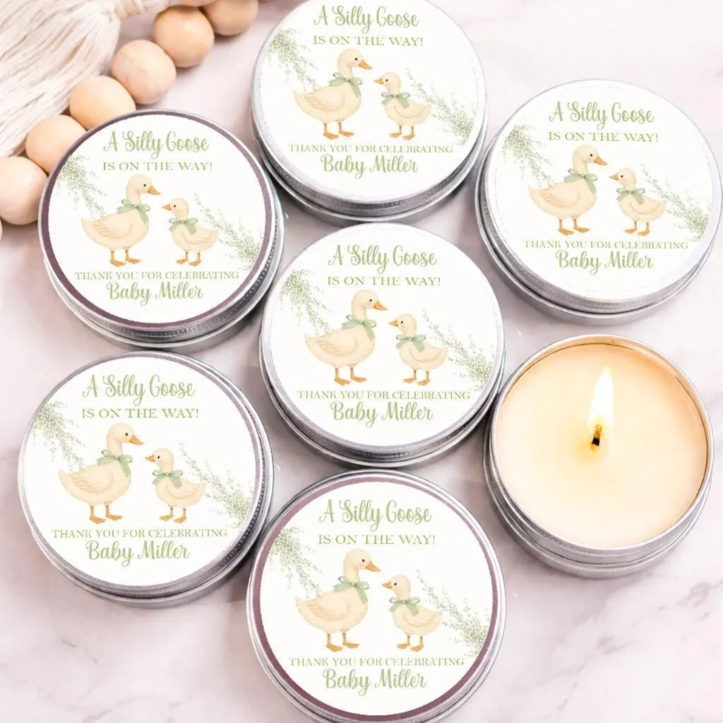 a collection of six tin candles with labels that read "A Silly Goose is on the way! Thank you for celebrating Baby Miller". The candles are arranged on a marble surface, with some of them lit, creating a warm and cozy atmosphere.