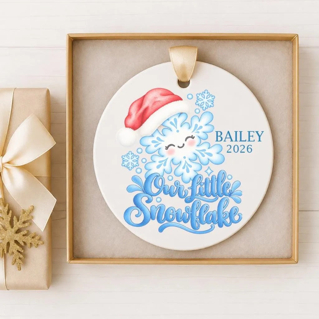 A white ceramic ornament with a red Santa hat and snowflakes, along with the text "Our Little Snowflake" and the name "BAiley" and the year "2026".