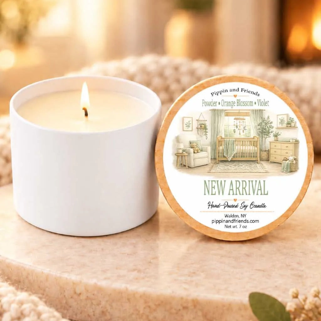 a white candle with a label that reads "New Arrival" and "Pippin and Friends" on a wooden surface. The candle is placed next to a white candle holder.