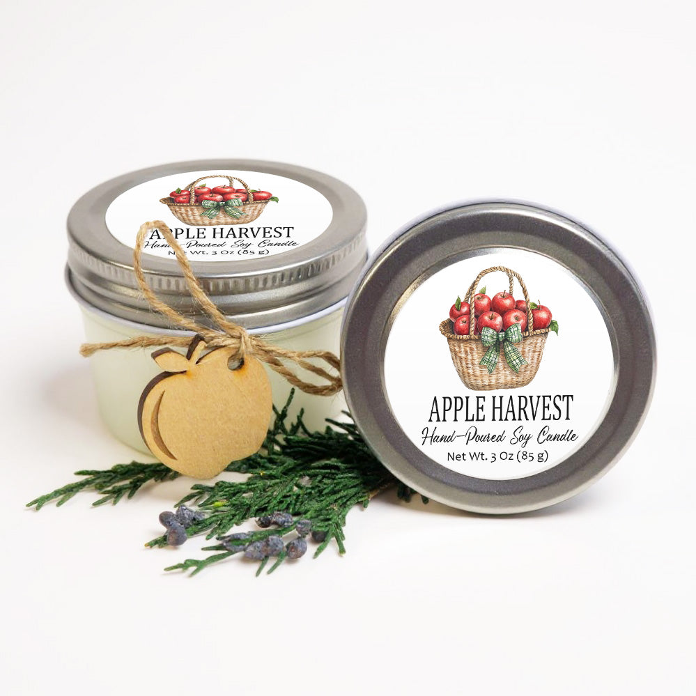 two jars of apple harvest soy candles, with a wooden tag attached to one of them. The jars are placed on a white background, and there are some pine branches and lavender sprigs nearby.