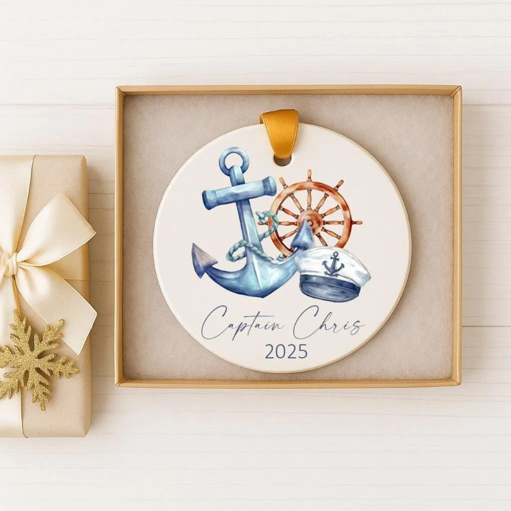 A decorative ornament featuring an anchor, a ship's wheel, and a captain's hat, with the text "Captain Chris" and the year "2025" printed on it. The ornament is displayed in a wooden box, and there is a gift box with a gold ribbon next to it.
