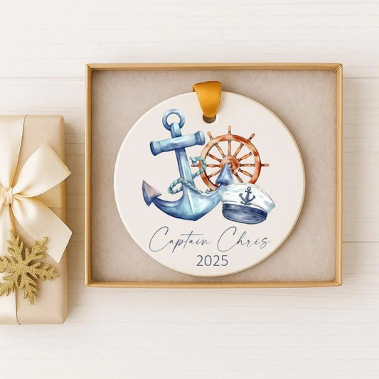 A decorative ornament featuring an anchor, a ship's wheel, and a captain's hat, with the text "Captain Chris" and the year "2025" printed on it. The ornament is displayed in a wooden box, and there is a gift box with a gold ribbon next to it.