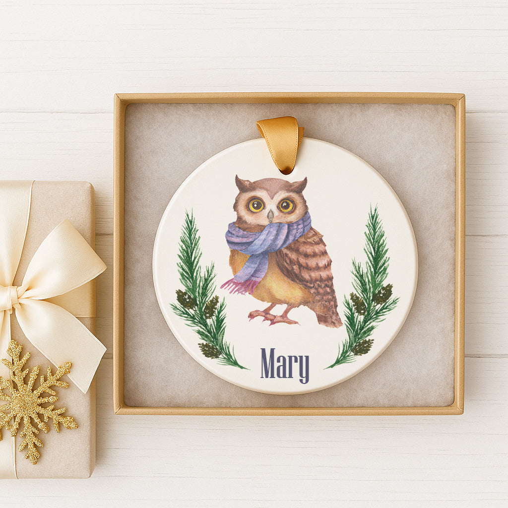 A ceramic ornament featuring an owl wearing a scarf and surrounded by pine branches, with the name "Mary" written on it. The ornament is displayed in a gold frame, and there is a gift box with a gold bow next to it.