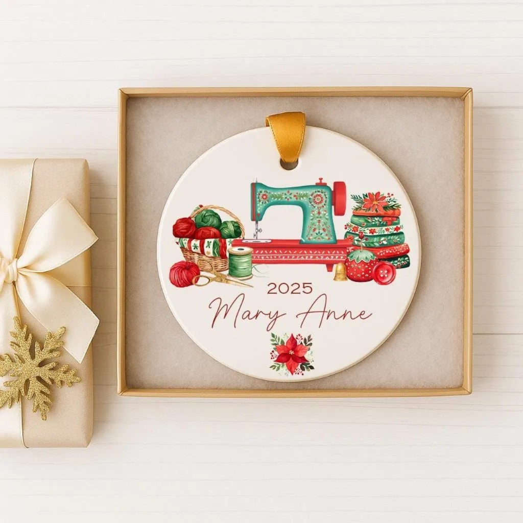 A ceramic ornament in a gold box, featuring a sewing machine surrounded by Christmas-themed items such as yarn, a red bow, and a red poinsettia flower.