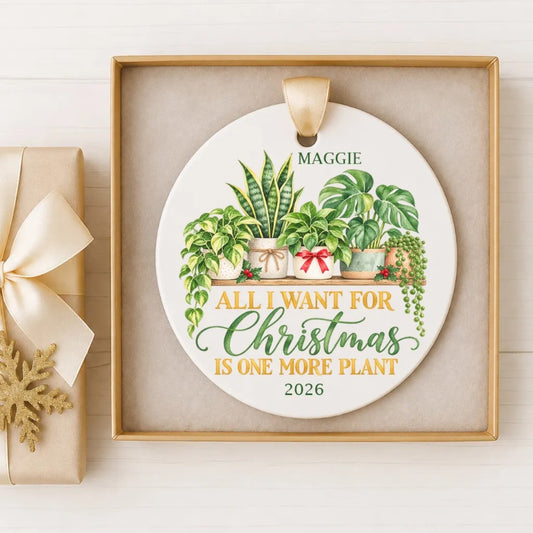 A white ceramic ornament with a gold frame, featuring a potted plant and the text "All I want for Christmas is one more plant" along with the year "2026".