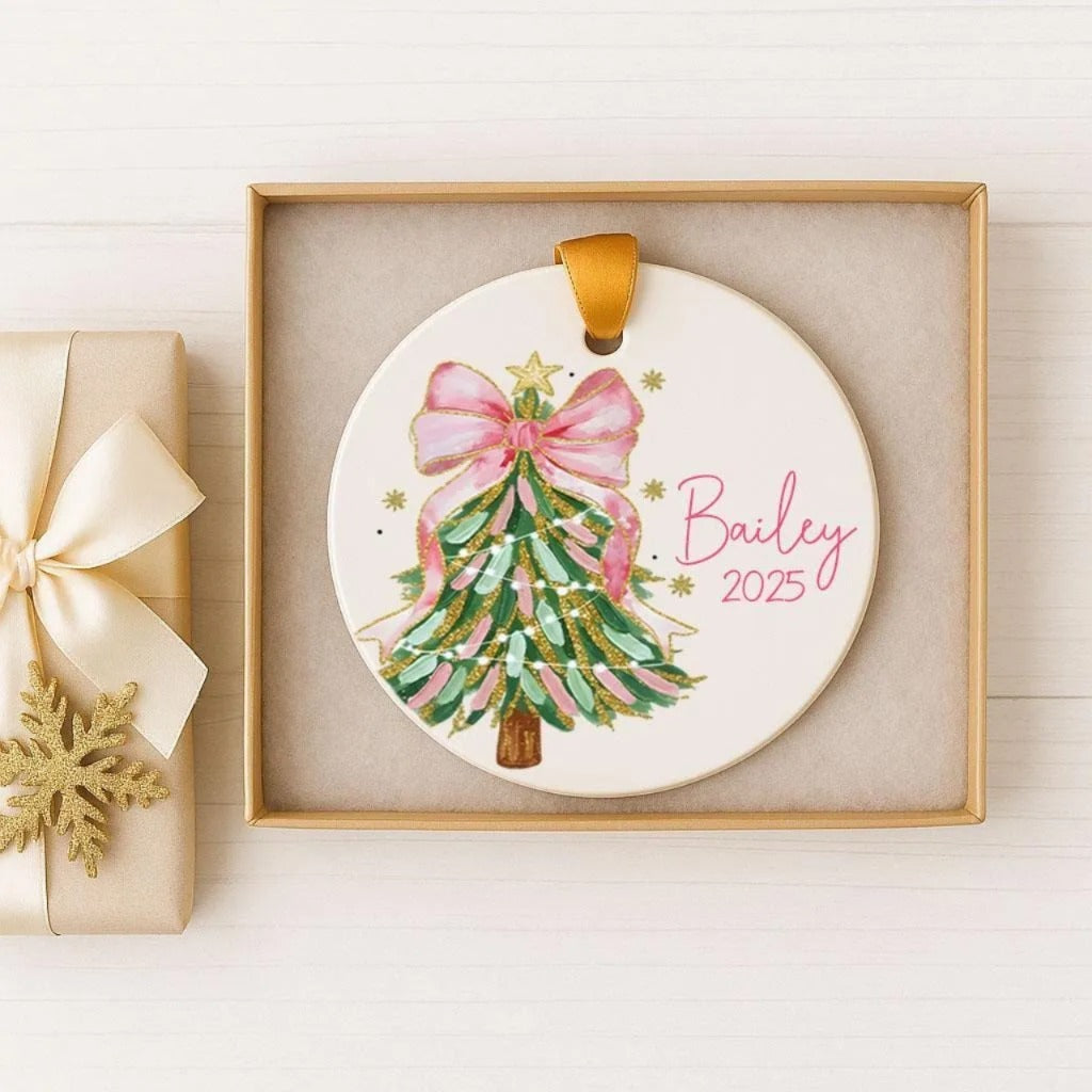 A white Christmas ornament with a pink bow and green and gold accents, displayed in a gold box.