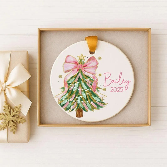 A white Christmas ornament with a pink bow and green and gold accents, displayed in a gold box.