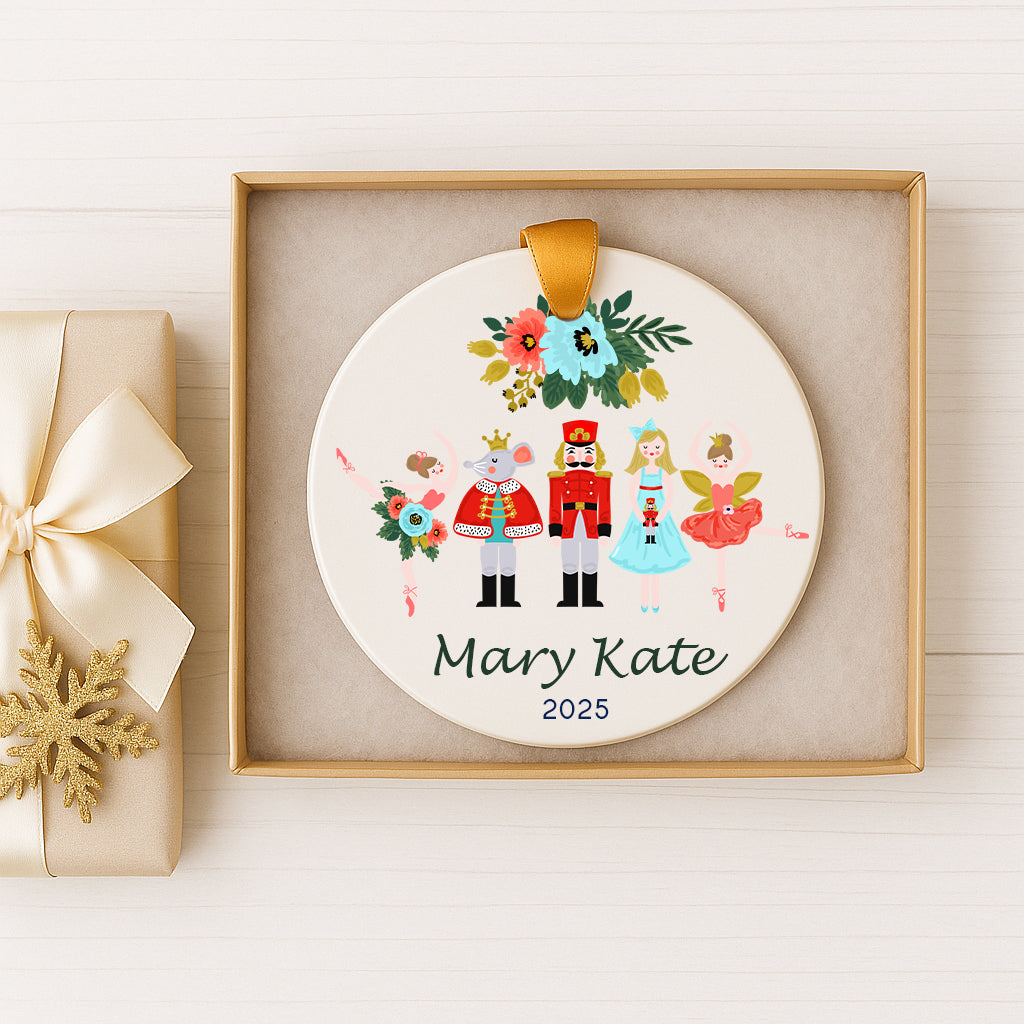A decorative ornament featuring a nutcracker, a king, and two fairies, with the name "Mary Kate" and the year "2025" printed on it. The ornament is displayed in a gold box, and there is a gift box with a gold ribbon next to it.