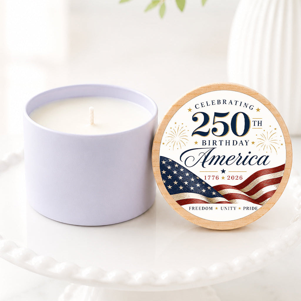 250th Birthday candle featuring a patriotic America-themed lid design with red, white, and blue flag artwork, displayed on a white pedestal cake stand. The candle is shown with the lid propped beside it in a bright, airy setting with soft greenery and white florals in the background. Ideal for America's 250th birthday celebration, patriotic party favors, and commemorative gifts
