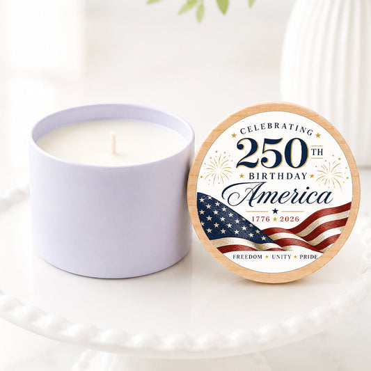 250th Birthday candle featuring a patriotic America-themed lid design with red, white, and blue flag artwork, displayed on a white pedestal cake stand. The candle is shown with the lid propped beside it in a bright, airy setting with soft greenery and white florals in the background. Ideal for America's 250th birthday celebration, patriotic party favors, and commemorative gifts