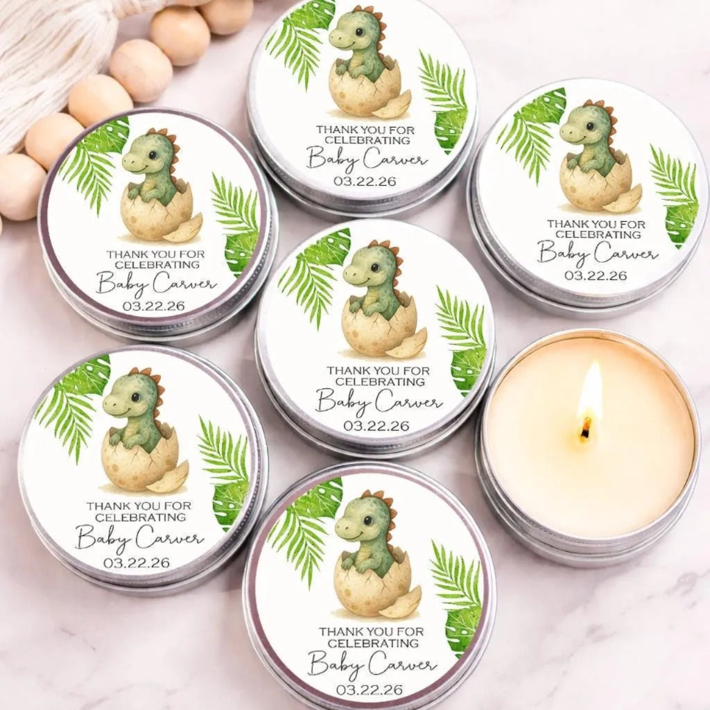 Group of personalized dinosaur baby shower candle favors displayed in silver tins on a marble surface. Each candle features a cute baby dinosaur hatching from an egg with tropical green leaves and custom text reading "Thank You for Celebrating Baby Carver." One open candle reveals creamy soy wax, while decorative wooden beads and a tassel accent the background.