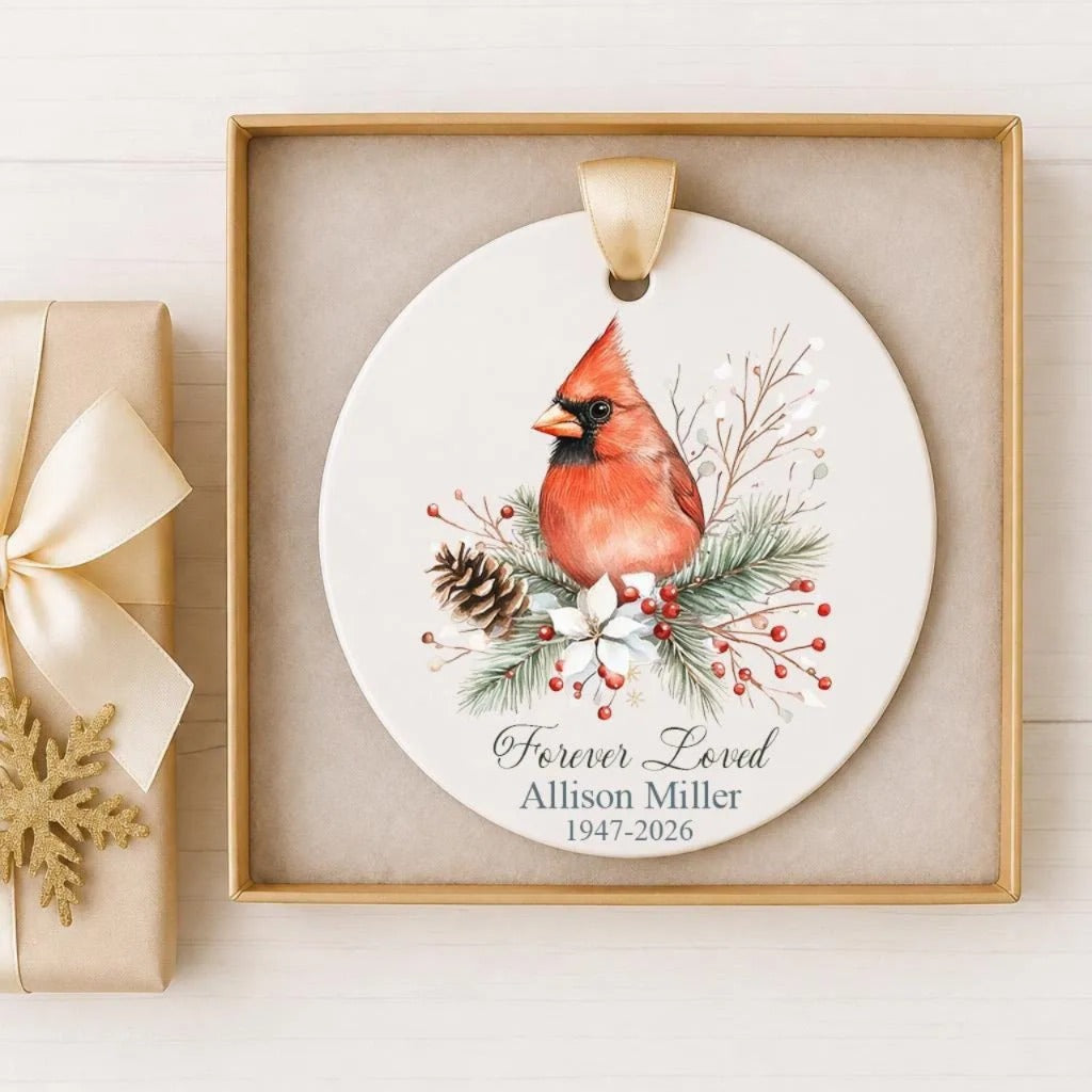 A white ceramic ornament with a red cardinal bird sitting on pine branches and berries, surrounded by a ribbon and a gold bow. The ornament is displayed in a gold frame.