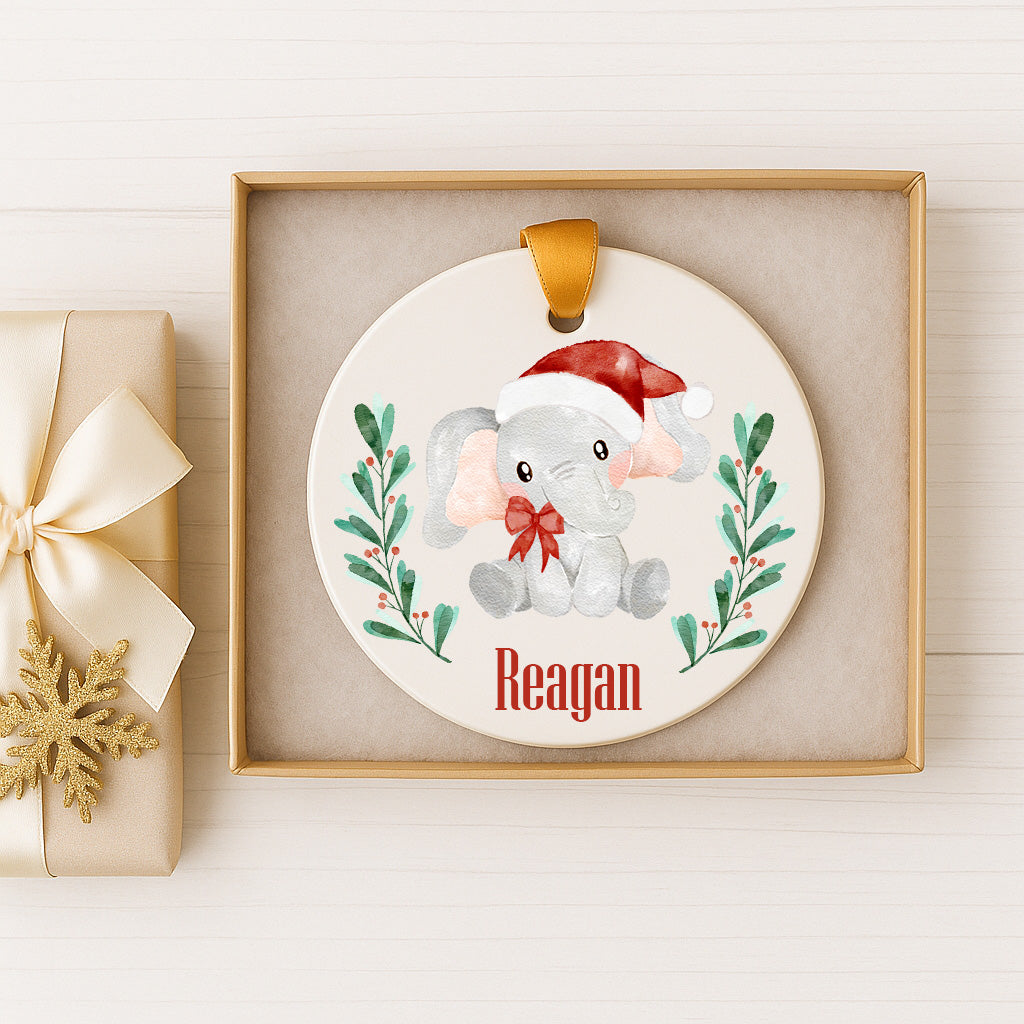 A white ornament with a red bow and a small elephant wearing a Santa hat, sitting on a green plant with red berries, is displayed in a gold box.