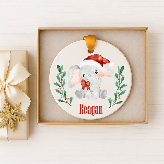 A white ornament with a red bow and a small elephant wearing a Santa hat, sitting on a green plant with red berries, is displayed in a gold box.