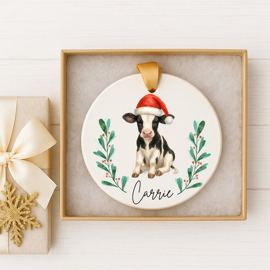 A ceramic ornament featuring a cow wearing a Santa hat, surrounded by green leaves and berries, with the name "Carrie" written on it. The ornament is displayed in a gold frame, and there is a gift box with a gold bow next to it.