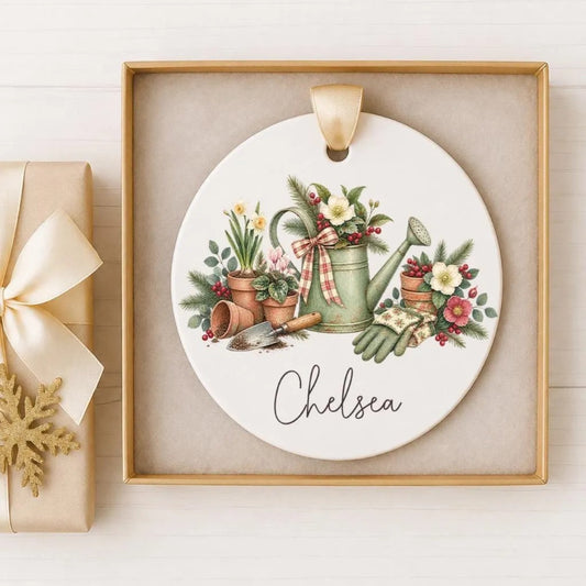 A decorative ceramic ornament featuring a garden scene with various plants, flowers, and gardening tools. The name "Chelsea" is written on the ornament.