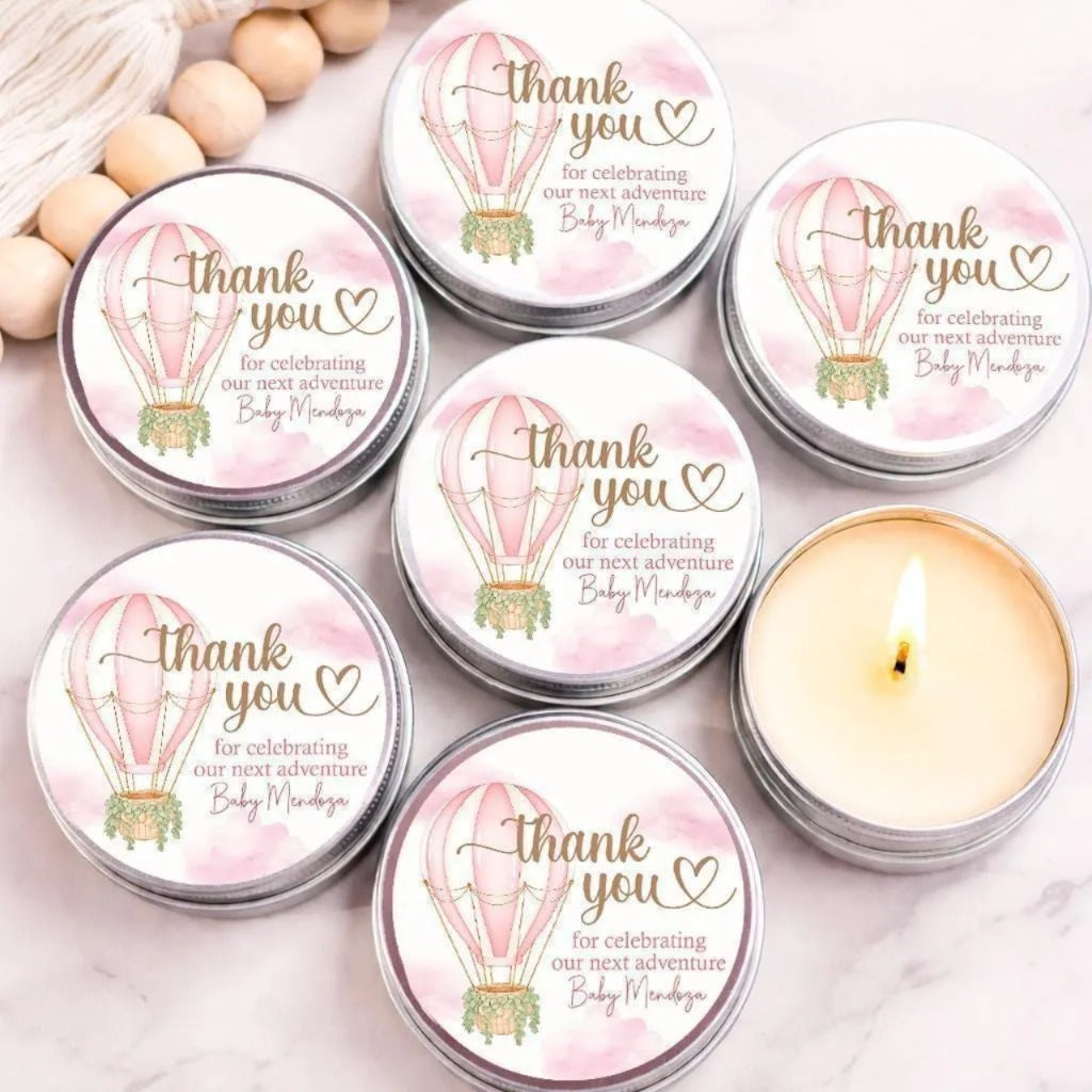 a collection of round tin containers with a pink and white color scheme, each featuring a heart and an illustration of a hot air balloon. The containers are arranged on a marble surface, with a lit candle in one of them. The containers are labeled with a message that reads "Thank you for celebrating our next adventure Baby Mendosa".