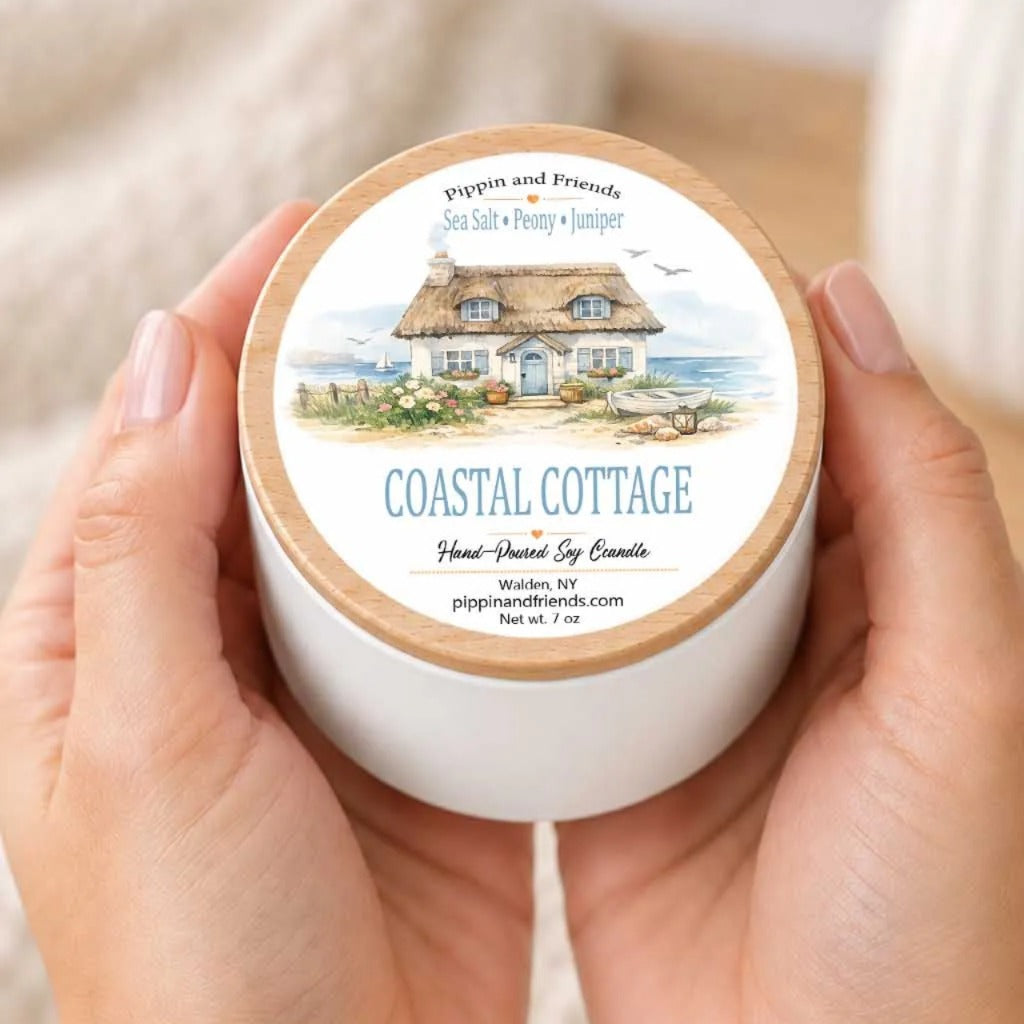 A person's hands holding a white container with a label that features an illustration of a coastal cottage.