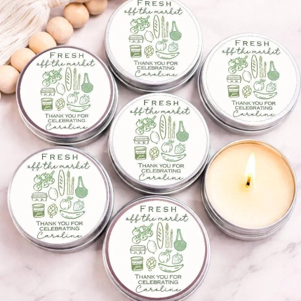 six silver tin candles with white labels, arranged on a marble surface. The labels feature illustrations of various food items and text that reads "Fresh off the market Thank you for celebrating Caroline".