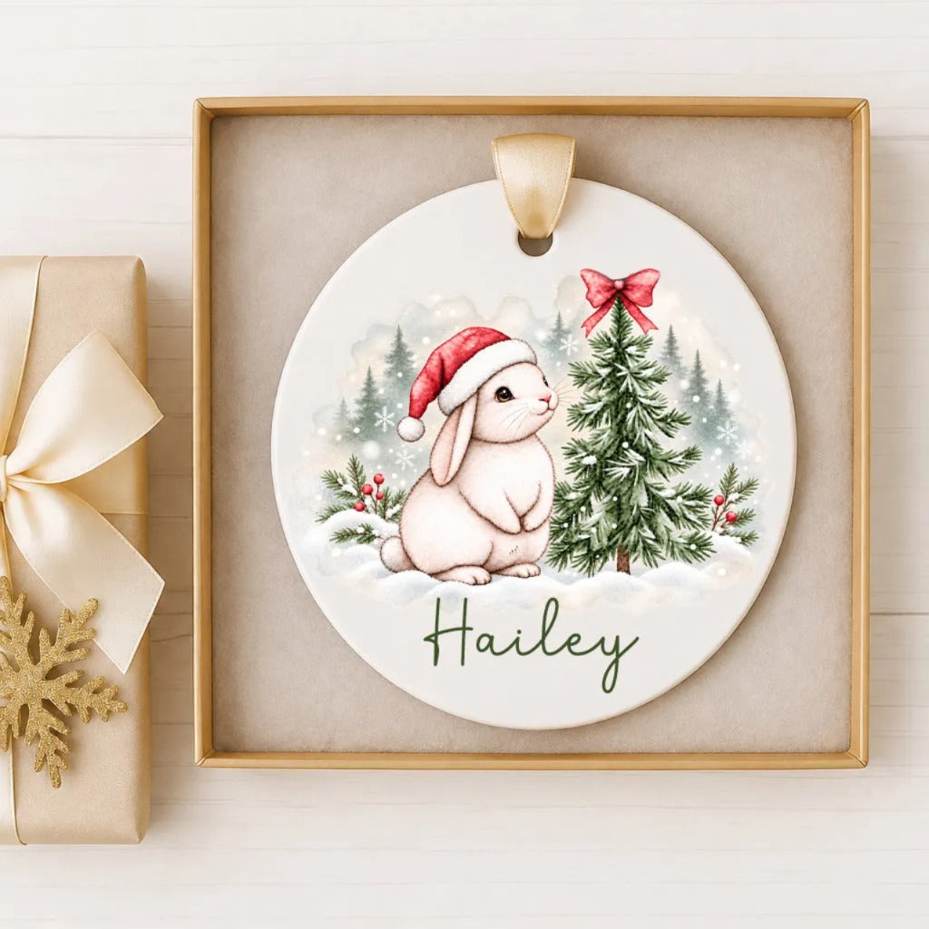 A ceramic ornament featuring a white rabbit wearing a Santa hat, sitting next to a Christmas tree, with the name "Haley" written on it. The ornament is displayed in a wooden frame, and there is a gift box with a gold bow next to it.