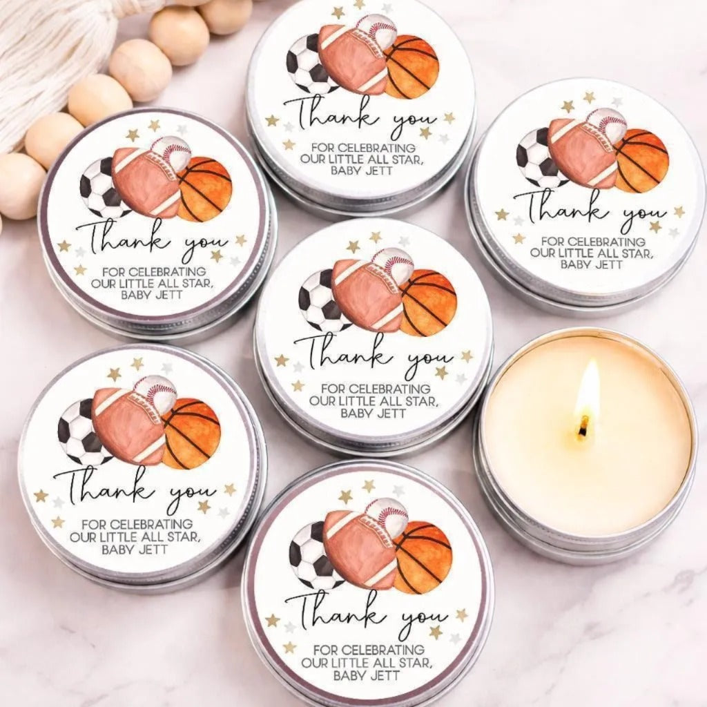 a collection of round tin candles with white labels, each featuring illustrations of sports balls and stars, along with a message that says "Thank you for celebrating our little all star, baby Jett".