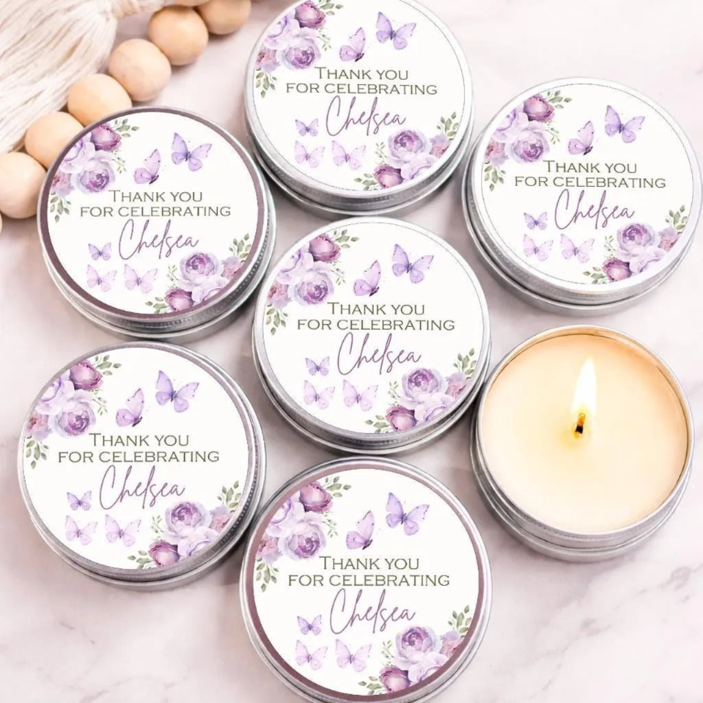 Set of personalized purple butterfly candle favors with watercolor floral labels in silver tins, displayed on a marble background with one candle lit.