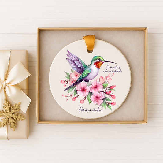A ceramic ornament featuring a colorful hummingbird surrounded by pink flowers, placed in a gold box.