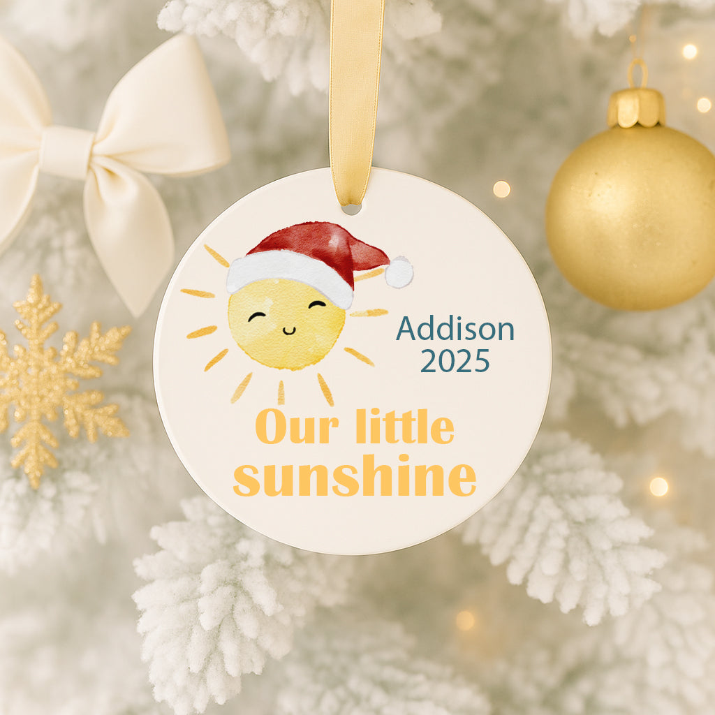 A white ornament with a smiling sun in a Santa hat, hanging on a Christmas tree.