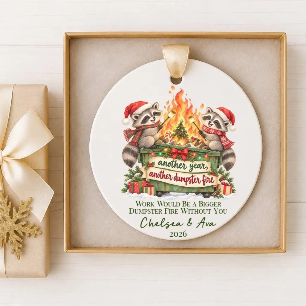 A white ceramic ornament with a festive design featuring two raccoons wearing Santa hats and scarves, sitting next to a fireplace with a sign that says "Another year, another dumpster fire". The ornament also includes text that says "Work would be a bigger fire without you Chelsea & Ava".