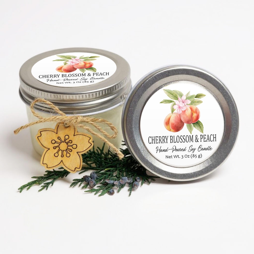 two jars of cherry blossom and peach scented soy candles, with a wooden flower charm and pine branches in the foreground.
