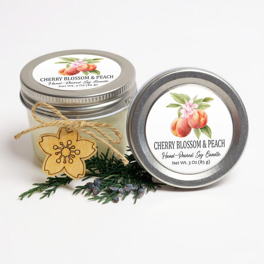 two jars of cherry blossom and peach scented soy candles, with a wooden flower charm and pine branches in the foreground.