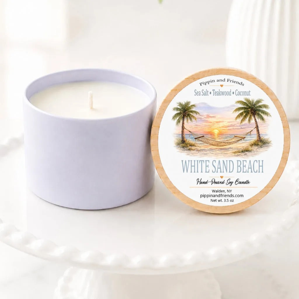 A soy candle in a matching cup sits beside its round lid on a white pedestal. The lid features a tropical beach illustration with palm trees and the text 'White Sand Beach' and 'Pipkin and Friends'.