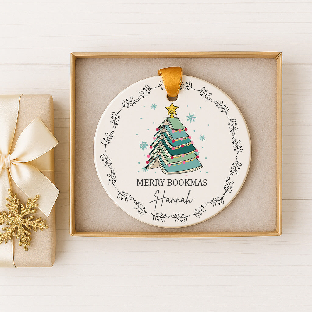 A white Christmas ornament with a green Christmas tree and snowflakes, along with the text "MERRY BOOKMAS Hannah" is displayed in a gold box.