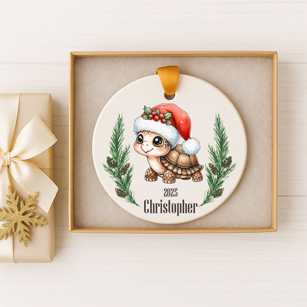 A ceramic ornament featuring a cartoon turtle wearing a Santa hat, surrounded by pine branches and a snowflake, with the name "Christoph" written on it. The ornament is displayed in a gold box, and there is a gift box with a gold ribbon next to it.