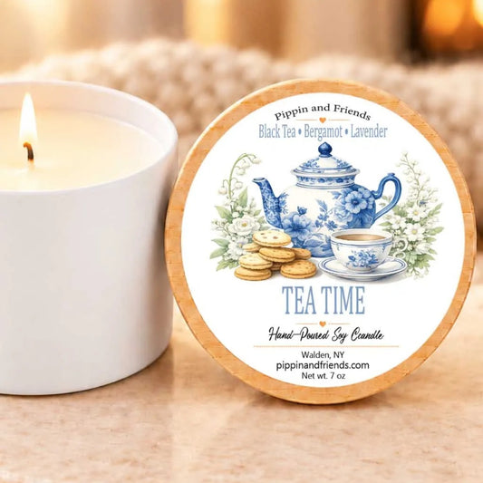 A white candle with a label that reads "Tea Time Hand-Drawn Soy Candle" next to a white candle holder.