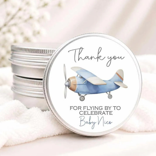 A round tin lid with a silver lid and a blue airplane illustration on it. The lid has a message that says "Thank you for flying by to celebrate Baby Nico".