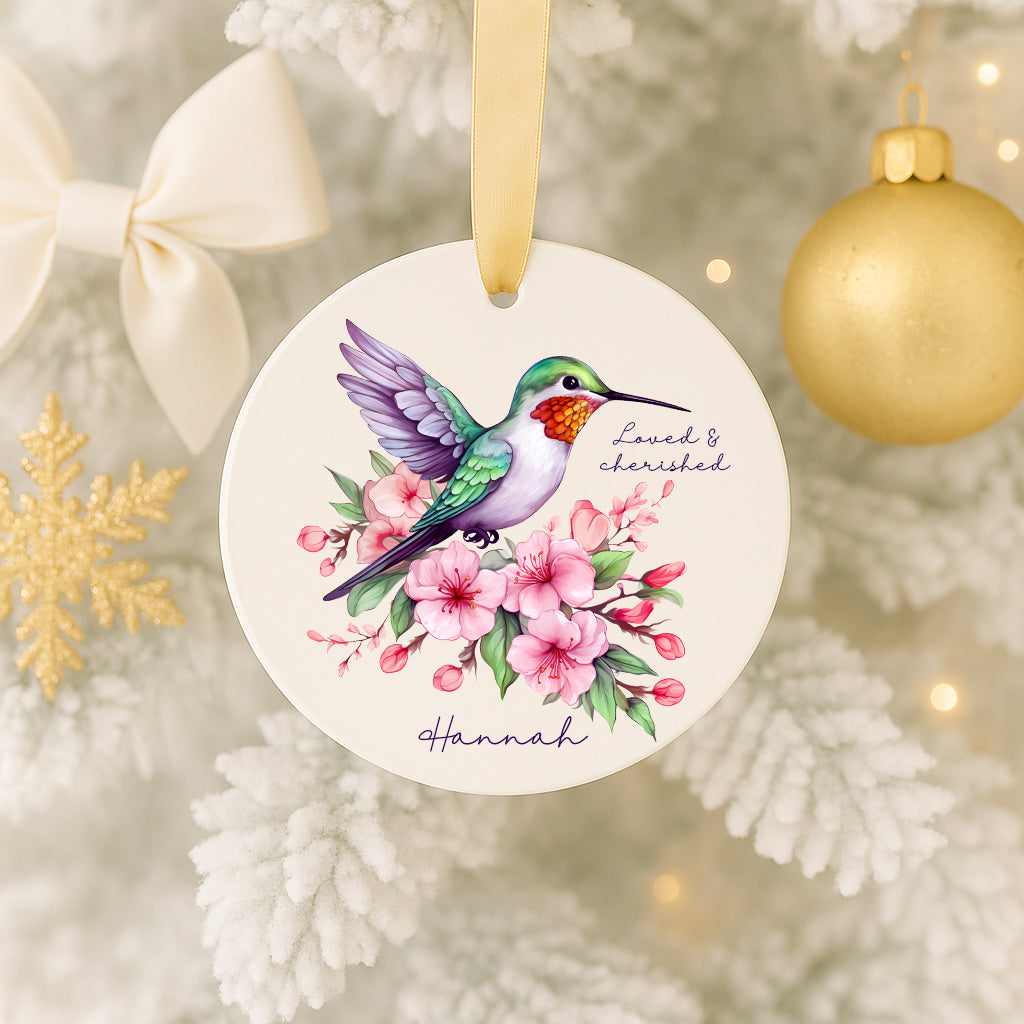 A white ornament with a colorful bird, pink flowers, and a ribbon is hanging on a Christmas tree.