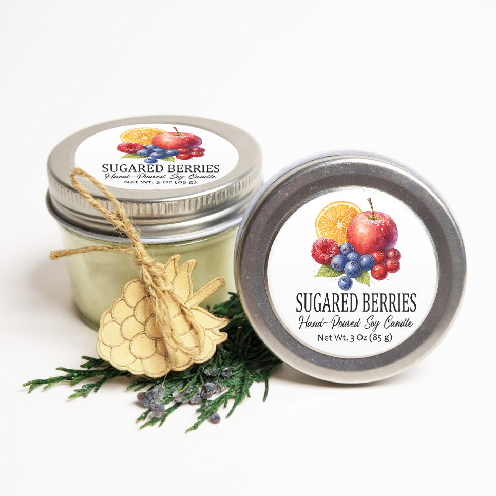 two jars of hand-poured soy candles, each adorned with a label that showcases a variety of fruits such as berries, apples, and oranges. The jars are placed on a white background, with a pine cone decoration and some greenery visible in the foreground.