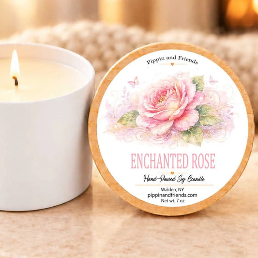 A white candle with a label that reads "Enchanted Rose" sits next to a lit candle on a table.