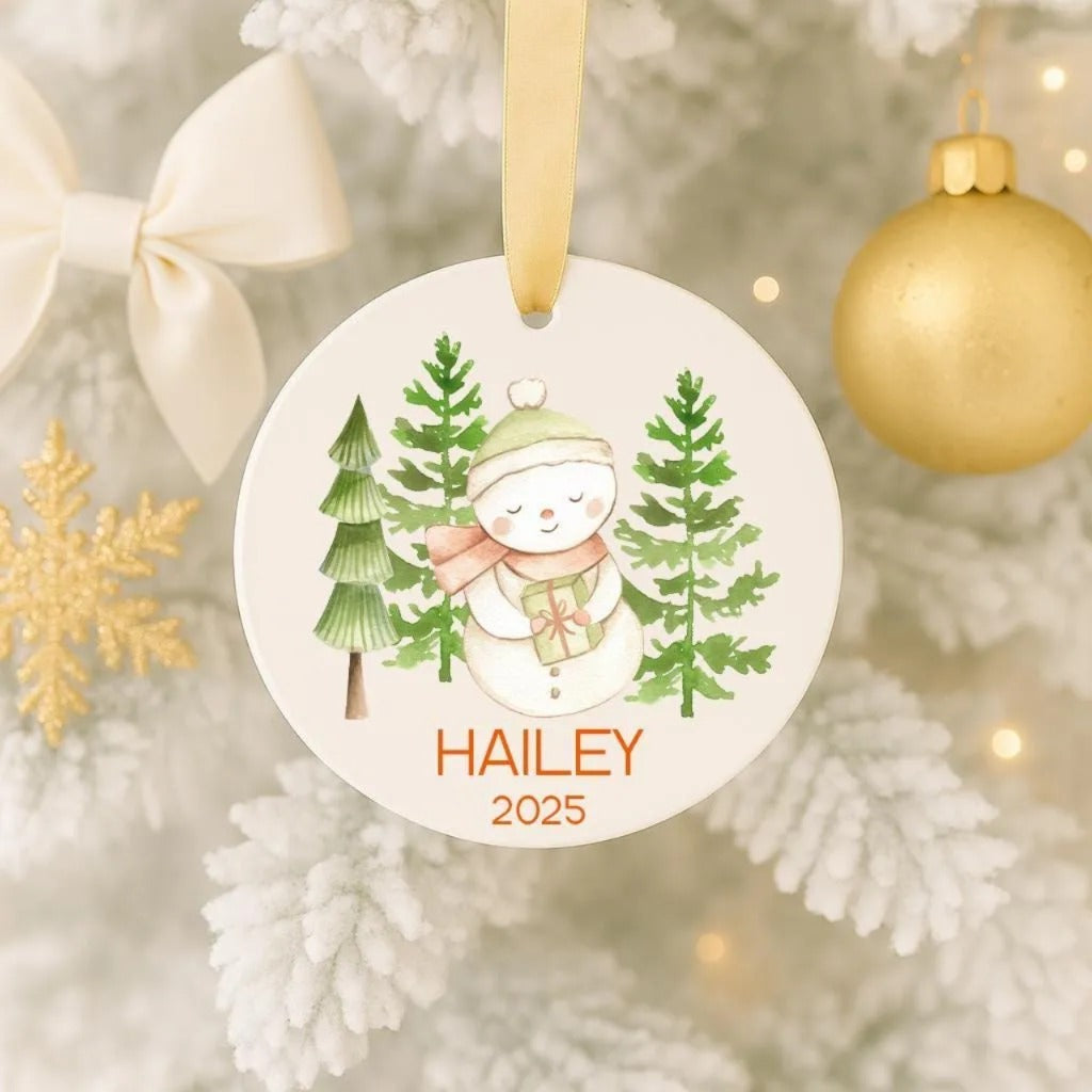 A white Christmas ornament with a snowman and pine trees, hanging on a tree.