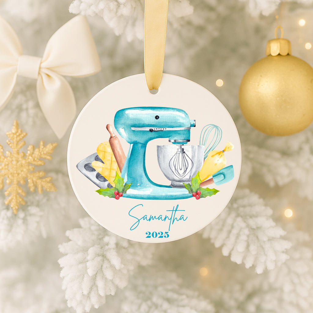 A Christmas ornament featuring a blue mixer surrounded by kitchen utensils, including a whisk, rolling pin, and spatula, with a snowflake ornament to the left.