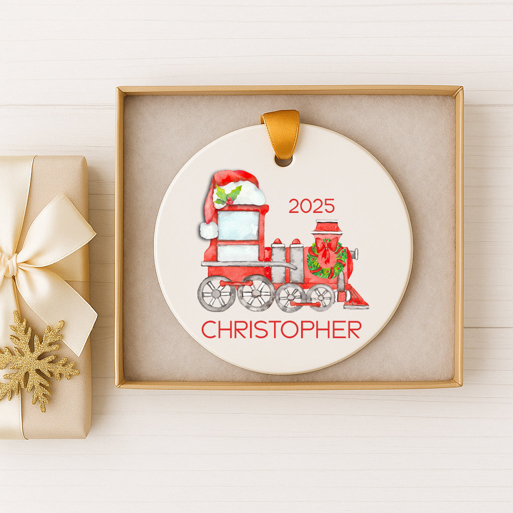 A Christmas ornament in a gold box, featuring a red and white train with a Santa hat and the name "CHRISTOPHER" written on it.
