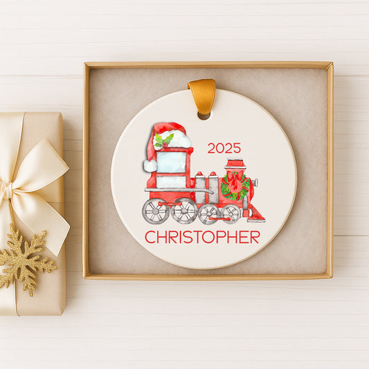 A Christmas ornament in a gold box, featuring a red and white train with a Santa hat and the name "CHRISTOPHER" written on it.