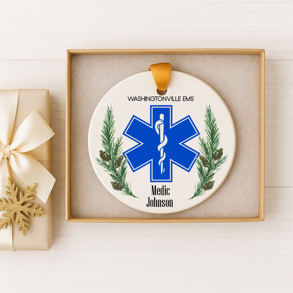 A white ornament with a blue star-shaped symbol and pine branches, featuring the text "Washingtonville Ems" and "Medic Johnson" on it. The ornament is displayed in a gold box, and there is a gift box with a gold ribbon next to it.