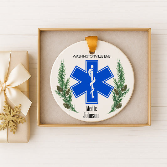 A white ornament with a blue star-shaped symbol and pine branches, featuring the text "Washingtonville Ems" and "Medic Johnson" on it. The ornament is displayed in a gold box, and there is a gift box with a gold ribbon next to it.