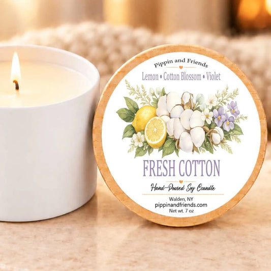 A white candle with a label that reads "Fresh Cotton" next to it, along with a blurred background that includes a candle and some flowers.